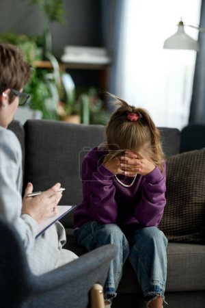Upset little girl sitting on sofa in bad mood while psychologist talking to her during their meeting at home