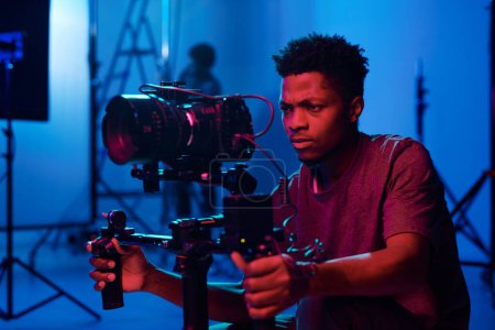 Foto de Joven operador de cámara afroamericano filmando en cámara profesional en estudio oscuro - Imagen libre de derechos