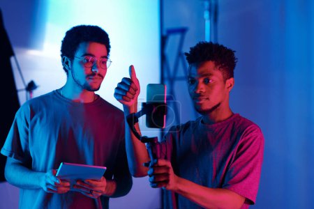Foto de Joven videomaker afroamericano usando smartphone para hacer contenido durante el trabajo en equipo con su colega en el estudio - Imagen libre de derechos