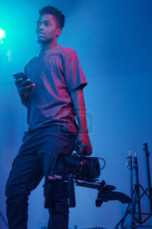 Foto de Operador de video afroamericano de pie en estudio oscuro con smartphone y cámara profesional - Imagen libre de derechos