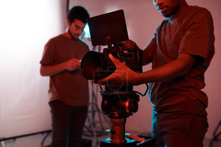 Foto de Joven operador de vídeo de pie delante de la cámara profesional y hacer película con su colega que trabaja en la tableta PC en segundo plano - Imagen libre de derechos