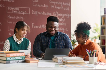 Foto de Grupo de tres jóvenes negros sonriendo felizmente mientras hacen el trabajo escolar juntos en la clase universitaria y mirando la pantalla del portátil - Imagen libre de derechos