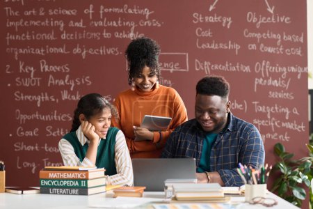 Téléchargez les photos : Vue de face portrait de trois étudiants noirs de collège regardant l'écran d'ordinateur portable contre tableau noir avec des écrits en arrière-plan - en image libre de droit