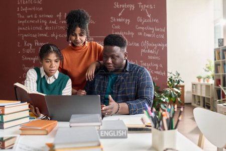 Téléchargez les photos : Portrait frontal de trois jeunes Noirs utilisant un ordinateur portable ensemble tout en travaillant sur un projet de groupe à l'école - en image libre de droit