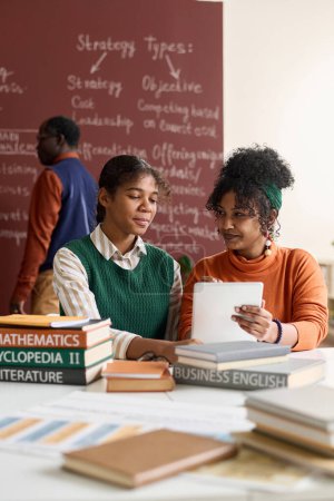 Téléchargez les photos : Portrait vertical de deux jeunes filles noires utilisant ensemble une tablette numérique pendant leurs études en classe - en image libre de droit
