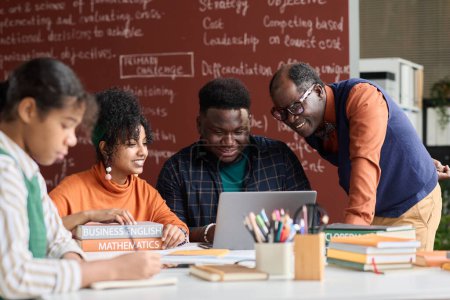 Téléchargez les photos : Professeur noir souriant aidant les élèves à travailler ensemble sur un projet de groupe et à utiliser un ordinateur portable - en image libre de droit