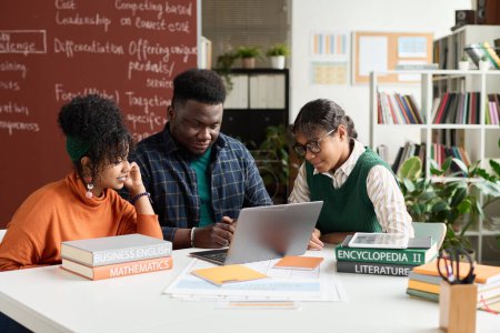 Téléchargez les photos : Groupe d'étudiants noirs utilisant un ordinateur portable dans leur étude pendant le travail d'équipe à table en classe - en image libre de droit