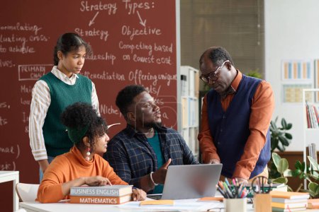 Téléchargez les photos : Portrait d'un enseignant afro-américain parlant à un groupe d'étudiants assis à table avec des livres et un ordinateur - en image libre de droit