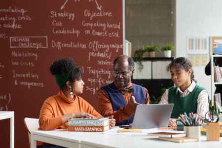 Téléchargez les photos : Portrait de l'enseignant noir senior parlant aux étudiants à la table dans la salle de classe du collège et pointant vers l'espace de copie d'écran d'ordinateur portable - en image libre de droit