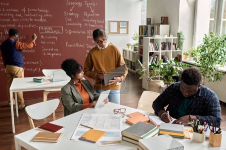 Téléchargez les photos : Vue en angle élevé chez les étudiants noirs faisant des études de groupe à table dans une salle de classe universitaire avec un espace de copie de tableau noir - en image libre de droit