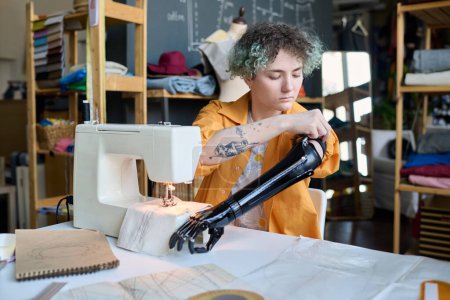 Photo for Portrait of teenage girl adjusting prosthetic arm while working in inclusive atelier studio and sewing clothing copy space - Royalty Free Image