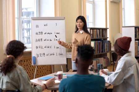 Portrait of young Asian woman teaching Chinese language class to group of students and pointing at hieroglyphs on whiteboard