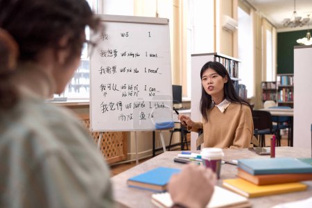 Portrait of young Asian woman as female teacher teaching Chinese language class sitting at table by whiteboard with hieroglyphs copy space