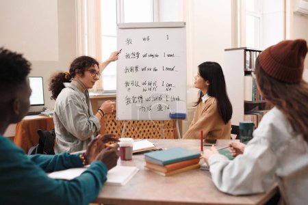 Retrato de vista lateral del estudiante masculino respondiendo preguntas durante la clase de idioma chino y apuntando a pizarra blanca con jeroglíficos