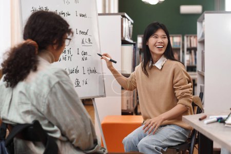 Portrait of young Asian woman laughing cheerfully pointing at whiteboard with hieroglyphs and enjoying fun Chinese language lesson copy space