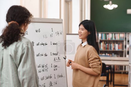 Retrato de una mujer asiática amable enseñando chino a un estudiante de pie junto a una pizarra en clase