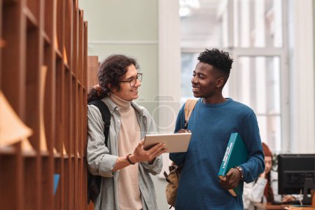 Retrato de la cintura hacia arriba de dos jóvenes sonrientes de pie junto a los estantes en la biblioteca de la universidad y utilizando espacio para copiar tabletas digitales