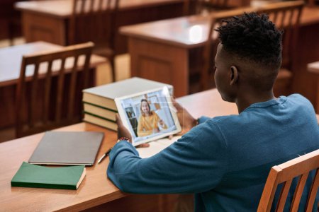 Über-die-Schulter-Ansicht eines jungen afroamerikanischen Studenten, der Videovorlesung in der Schulbibliothek anschaut und ein digitales Tablet nutzt, während der Lehrer auf dem Bildschirm spricht