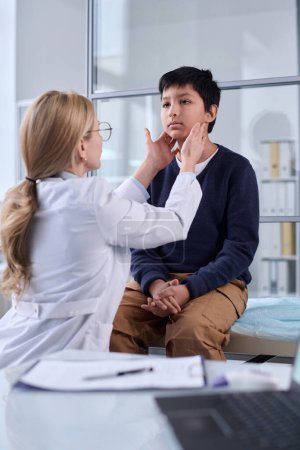 Vertical portrait of pediatrician examining neck and throat of young boy during health check up in clinic