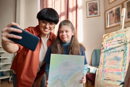 Retrato de la cintura hacia arriba de la joven sonriente con discapacidad tomando foto selfie con el profesor de arte en el estudio y sosteniendo la imagen