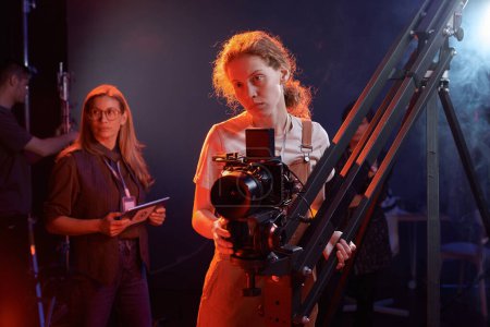Waist up portrait of camerawoman operating equipment on rig in studio with red neon lights copy space