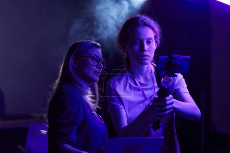 Tailliertes Porträt weiblicher Videoproduktionsteams, die zusammen arbeiten und das Smartphone in lila Neonlicht benutzen
