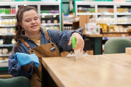 Waist up portrait of smiling young woman with disability working in supermarket and sanitizing tables in cafe area copy space