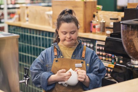 Waist up portrait of young woman with Down syndrome working in cafe and cleaning coffee cups enjoying occupational training
