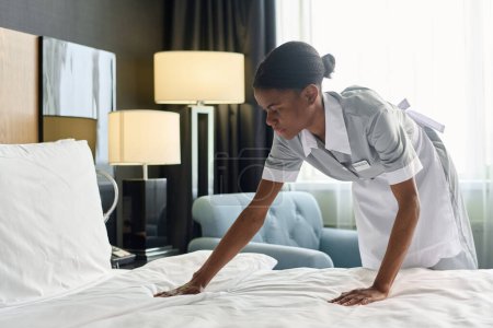 Side view portrait of young African American housekeeper working in hotel and making bed with fresh sheets copy space