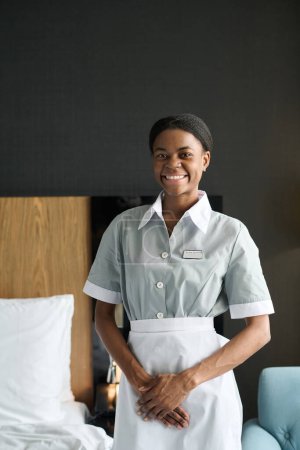 Retrato vertical de una mujer afroamericana sonriente como ama de llaves con uniforme mirando a la cámara en el dormitorio