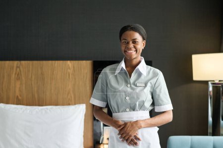 Waist up portrait of young Black woman as housekeeper wearing uniform and smiling at camera standing in hotel room copy space