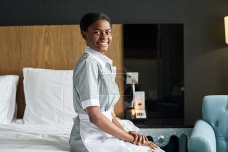 Retrato de vista lateral de la joven mujer negra como ama de llaves con uniforme y sonriendo a la cámara sentada en la cama en el espacio de copia de la habitación del hotel