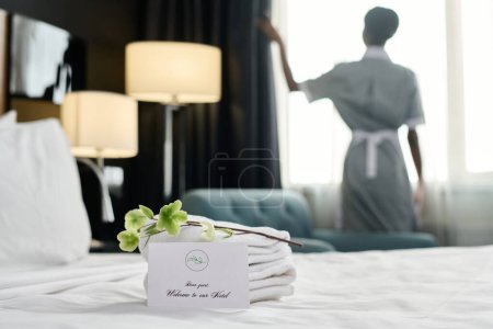 Welcome note with delicate flower decor on bed in luxury in hotel room and blurred silhouette of housekeeper copy space