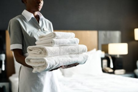 Recortado tiro de joven afroamericana mujer como ama de llaves celebración pila de ropa de cama fresca preparando habitación en el espacio de copia de hotel de lujo
