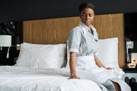 Retrato de una joven mujer negra trabajando como ama de llaves sentada en la cama en el espacio de copia de la habitación del hotel