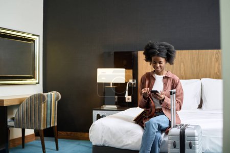 Wide angle view at young Black woman sitting on bed in hotel room and using smartphone with suitcase copy space