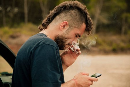 Junger Mann raucht Marihuana-Joint, während er sein Smartphone kontrolliert