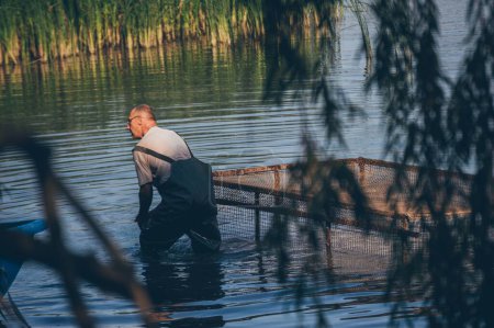 Téléchargez les photos : Pêcheur en caoutchouc et filet de pêche dans l'eau derrière le saule - en image libre de droit