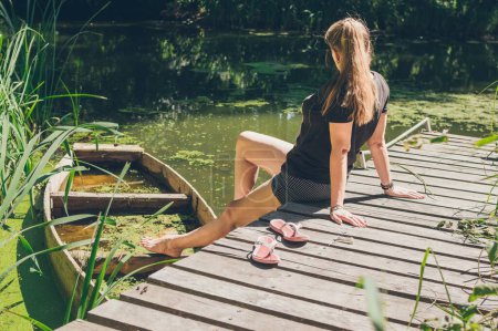 Téléchargez les photos : Femme assise au soleil sur un pont en bois par un vieux bateau - en image libre de droit