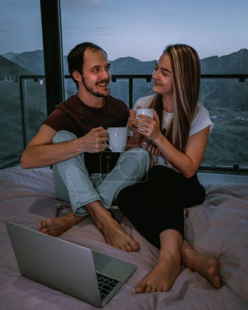 Foto de Bonita pareja en casa acogedora, sentada en la cama, bebiendo un té - Imagen libre de derechos