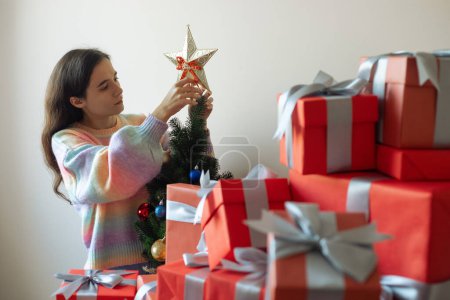 Foto de Una mujer morena en un suéter se aferra a una estrella en el árbol de Año Nuevo festivo. regalos rojos en primer plano - Imagen libre de derechos