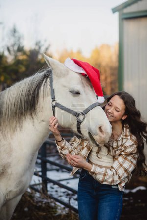 Téléchargez les photos : Cheval blanc avec chapeau de Père Noël et jeune fille souriante - en image libre de droit