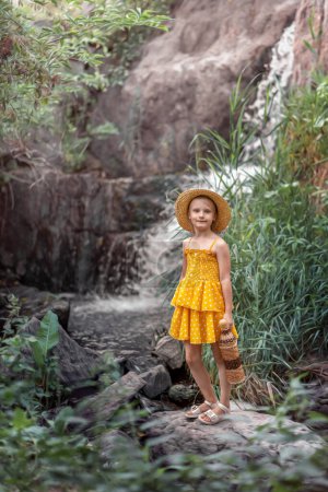 Photo for A girl in a hat and a yellow dress stands by a stream - Royalty Free Image