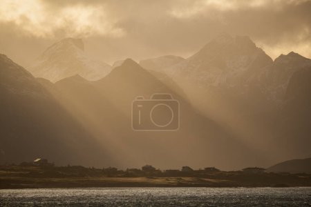 Téléchargez les photos : Rayons de lumière sur les maisons et les montagnes lointaines près de Ramberg, Flakstady, Lofoten Islands, Norvège - en image libre de droit