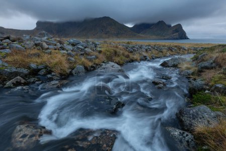 Foto de Pequeño río rocoso que fluye a través del paisaje herboso debajo de los picos de montaña, Unstad, Vestvgy, Islas Lofoten, Noruega - Imagen libre de derechos