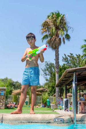 Téléchargez les photos : Garçon aux cheveux bruns jouant avec le pistolet à eau dans la piscine d'un camping dans un pays tropical - en image libre de droit