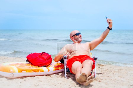 Téléchargez les photos : Homme chauve jouer et profiter de son flotteur et mobile, se baigner et prendre des selfies sur la plage - en image libre de droit