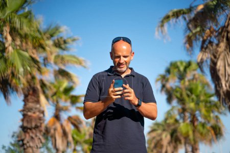 Foto de Hombre calvo hablando y usando su teléfono inteligente en un país tropical con sus palmeras - Imagen libre de derechos