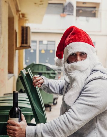 Foto de Santa Claus sin hogar bebiendo vino y buscando en los cubos de basura - Imagen libre de derechos