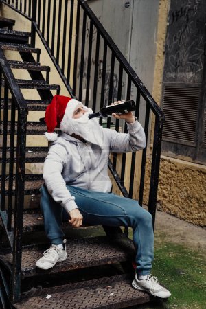 Foto de Santa Claus sin hogar bebiendo vino y buscando en los cubos de basura - Imagen libre de derechos